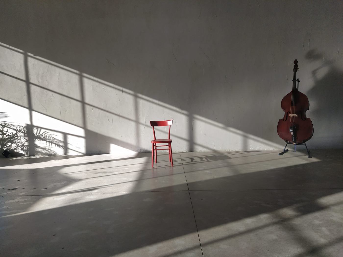 Una sedia rossa e un contrabbasso soli nella sala vuota, con l'ombra della vetrata disegnata sul pavimento in cemento.